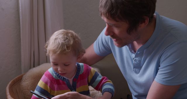 Father holding digital tablet while sitting with toddler in cozy home environment, both engaged with screen. Suitable for topics related to modern parenting, technology use among young children, family lifestyle, and digital learning. Ideal for use in blog posts, parenting articles, advertisements for family-oriented tech products, and educational resources.