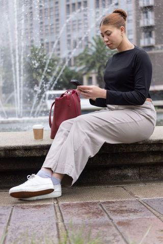 Young african american woman checking phone on fountain ledge with red handbag and coffee