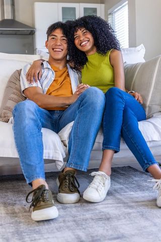 Happy diverse couple cuddling on couch, smiling and holding hands in bright living room