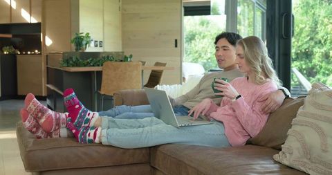 Couple relaxing on sectional sofa using laptop and drinking coffee in modern open-plan living room