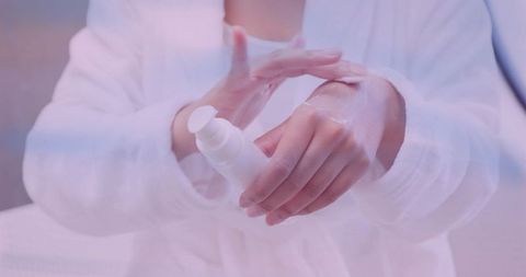 Indian woman applying hydrating hand lotion from pump bottle while wearing white bathrobe