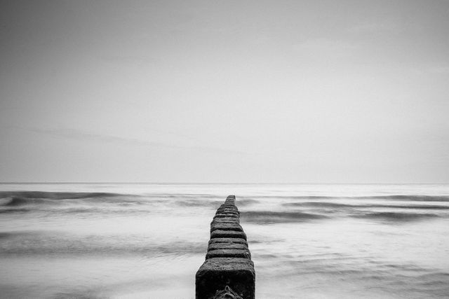 Monochrome photo shows long stone jetty stretching into ocean. Shot with long exposure, creating glassy water effect. Perfect for promoting relaxation, meditation apps, or travel blogs. Ideal addition to minimalist, modern interiors seeking soothing, natural imagery.