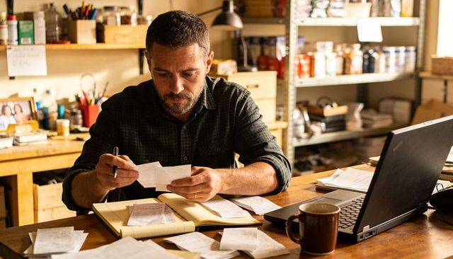 Man counting receipts at home workshop desk with laptop and notepad