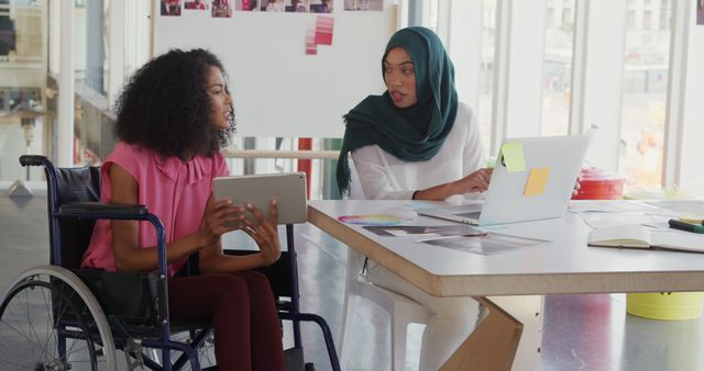Front view of two young biracial female business creatives working together and talking in a modern office, one is sitting in a wheelchair and holding a tablet computer, the other woman who is wearing a hijab is sitting at a desk using a laptop computer