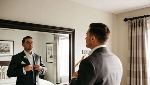 Groom adjusting jacket and boutonniere in mirror preparing for wedding ceremony