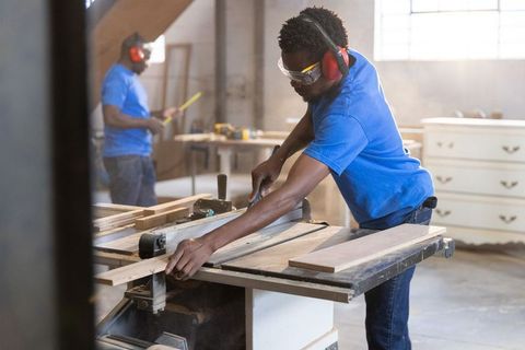 African american carpenter cutting wood on table saw in busy workshop showing teamwork
