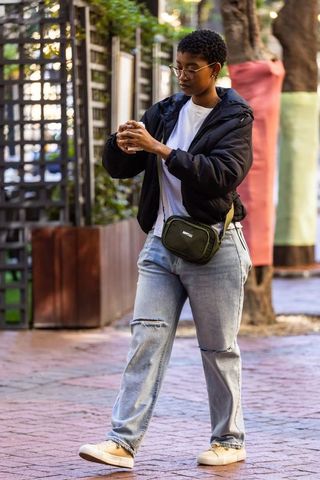 African american woman walking sidewalk checking phone wearing black puffer and olive crossbody