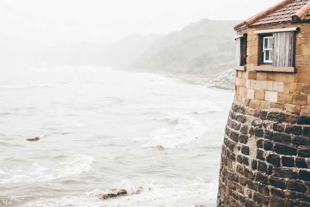 Rustic stone house perched on coastline, facing foggy ocean waves. Imagine this scene used for themes like coastal living, maritime charm, tranquil retreats, or nature's beauty. Ideal for travel brochures, inspiration boards, and background settings for blogs focusing on sea and coastal imagery.