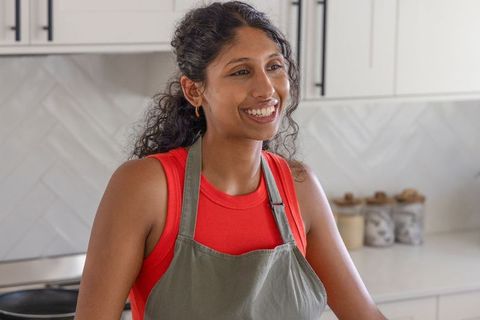 Indian woman smiling while cooking in modern kitchen wearing gray apron and red top