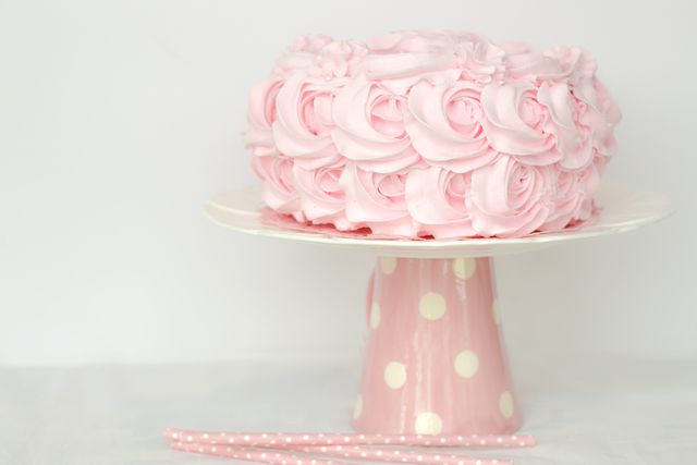 Decorative pink rose cake with intricate frosting design on a polka dot cake stand. Ideal for events like birthdays, weddings, or baby showers. Perfect for use in blogs, party invitations, or confectionery marketing materials highlighting elegant and visually appealing desserts.
