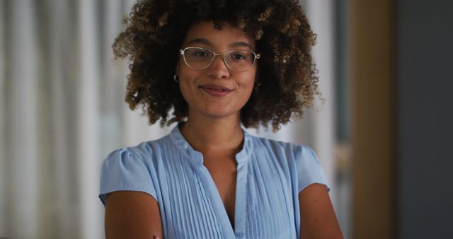 Young woman with curly afro hairstyle, dressed in blue blouse and glasses, smiling confidently with arms crossed indoors. Perfect for promotional material related to self-confidence, modern fashion, or eyewear advertisements.