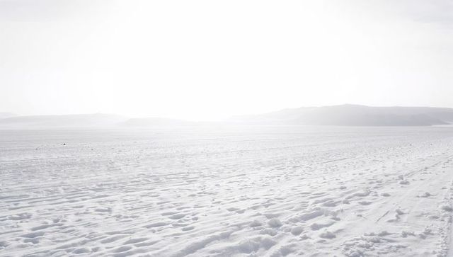 Minimalist frozen snow plain showing rippled ridges, tracks and distant hills at sunrise