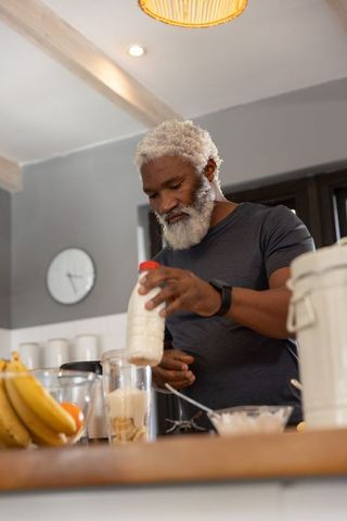 Senior african american man pouring milk and making banana smoothie in cozy kitchen