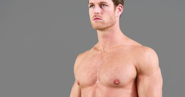 Athletic young man posing shirtless against gray background, showcasing his muscular physique and healthy body. Perfect for use in fitness, health, and motivational content, advertisements for gym memberships, and bodybuilding promotions. Ideal for illustrating strength, wellness, and dedication in training programs.