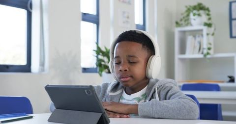 African american boy learning on tablet while wearing headphones in bright classroom