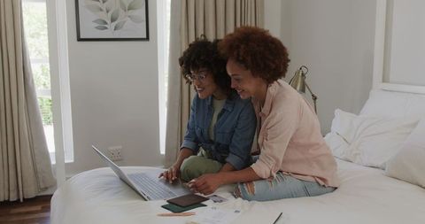 Two women using laptop on bed planning travel and finances in cozy sunlit bedroom
