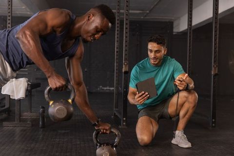 Asian trainer coaching African American client during kettlebell row workout with tablet, stopwatch