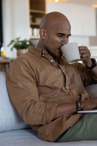 Middle-aged african american man sipping from white mug while reading tablet on sofa
