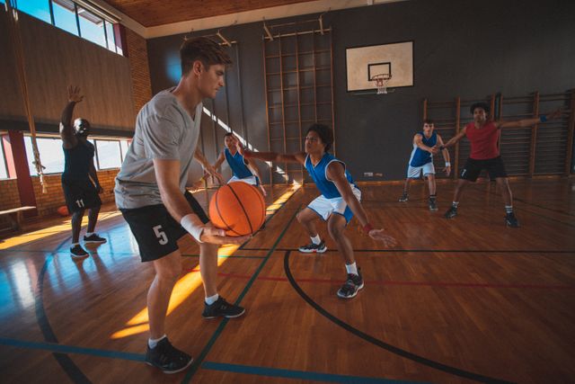 Diverse male basketball team practicing in a sunny indoor court with a coach raising his hand. Ideal for use in sports training programs, teamwork and strategy articles, fitness and health promotions, and advertisements for sports equipment and apparel.