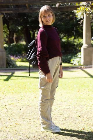 Young woman standing in sunlit park wearing maroon sweater and backpack casual outdoor portrait