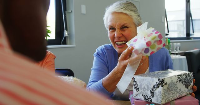 A senior Caucasian woman is joyfully receiving a gift from someone, with copy space. Her delighted expression suggests a moment of happiness and celebration, during a birthday or special occasion.