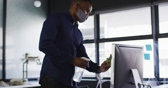 Man wearing face mask disinfecting an office desk with spray and cloth, emphasizing workplace hygiene and COVID-19 safety protocols. Ideal for corporate health and safety campaigns, articles on pandemic workplace practices, and educational materials on sanitation.