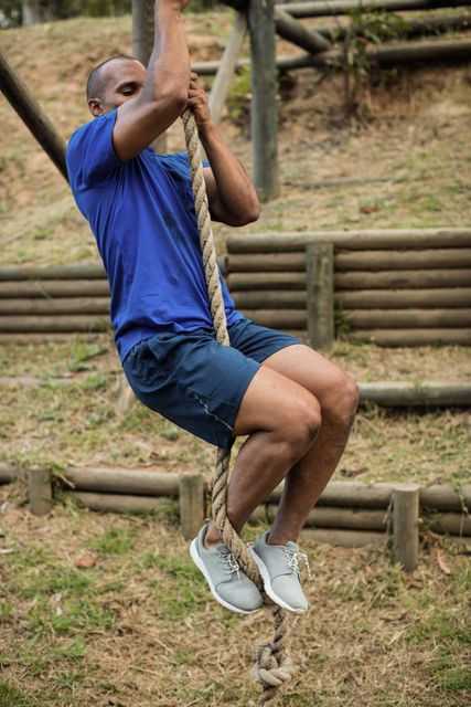 Fit man climbing rope during obstacle course in boot camp. Ideal for use in fitness and training materials, motivational posters, military training programs, and outdoor adventure promotions. Highlights themes of strength, determination, and physical challenge.
