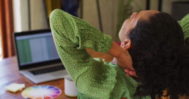 Woman with curly hair in green blouse stretching her neck while working from home on laptop. Useful for articles about ergonomics, home office productivity, break time during work, and staying healthy while working remotely.