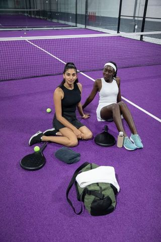 Diverse women sitting on purple paddle court wearing sporty black and white outfits with paddles