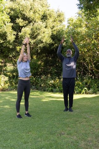 Mature couple stretching on sunny backyard lawn wearing fitness trackers