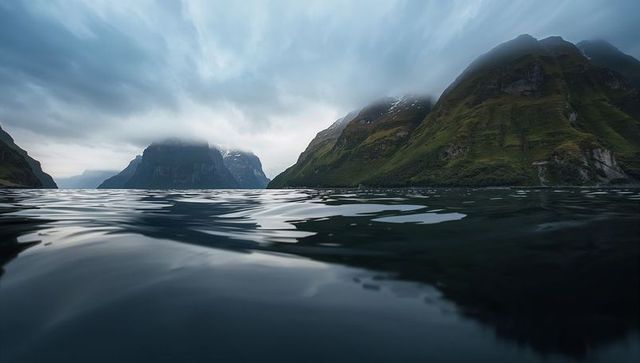 Glassy water reflecting fjord leading toward misty mountains and dramatic sky