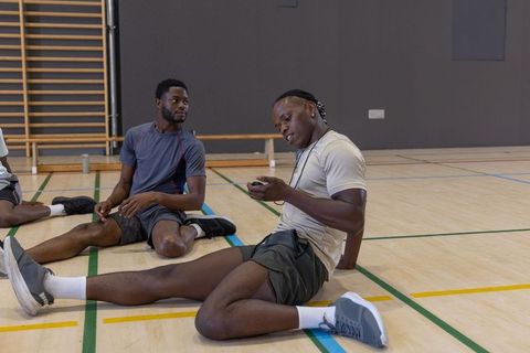 African american athletes sitting on gym court holding smartphone and stopwatch lanyard