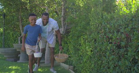 African american father and son hunting easter eggs in sunny backyard with woven baskets