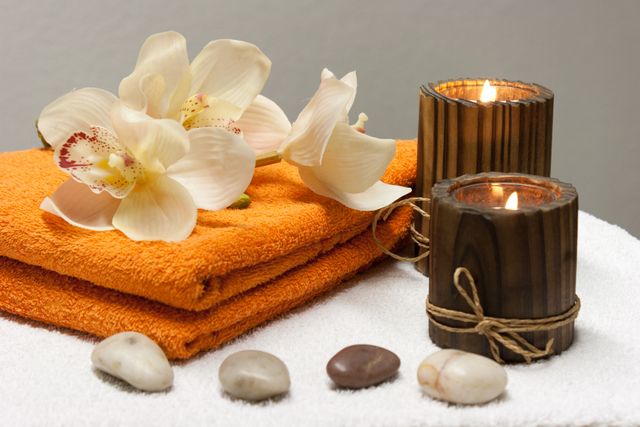 This serene image captures a relaxing spa setting with burning candles, neatly folded orange towels, and beautiful orchid flowers. Accompanying the setup are smooth stones, adding to the peaceful atmosphere. Perfect for use in wellness blogs, spa service advertisements, lifestyle articles, or relaxation-themed projects.