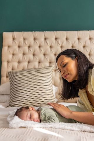 Mother leaning over sleeping baby on cozy bed with tufted headboard, gentle daylight moment