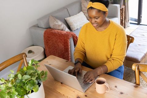 African american woman working from home at cozy sunlit table with laptop coffee plant