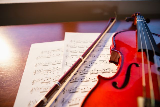 This image shows a violin and bow placed on top of sheet music on a wooden table, illuminated by sunlight. Ideal for use in educational materials, music school promotions, classical music event flyers, and articles about music education or learning to play instruments.