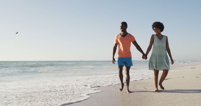 Smiling african american couple holding hands and walking on sunny beach. healthy, active family beach holiday.