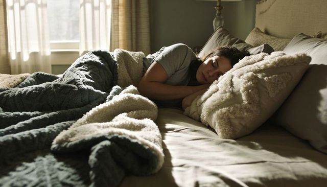 Sleeping woman lying on side under plush blanket in sunlit cozy bedroom for relaxation