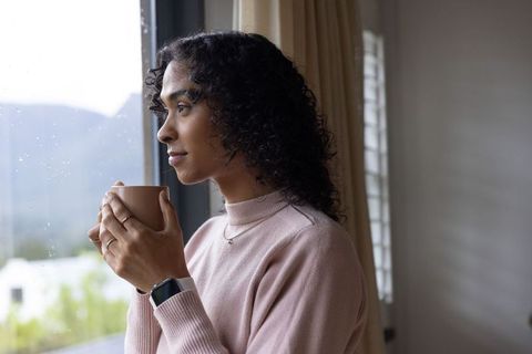 Non-binary adult sipping warm mug by rain-speckled window wearing pink sweater