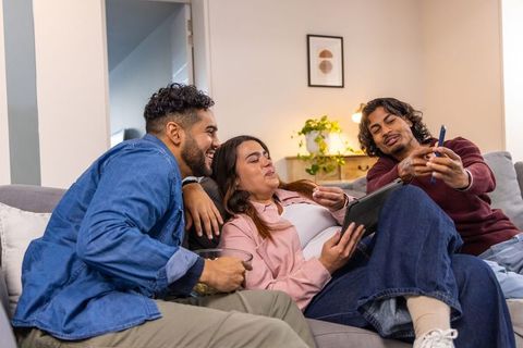Asian friends laughing and sharing tablet while relaxing on gray couch at home