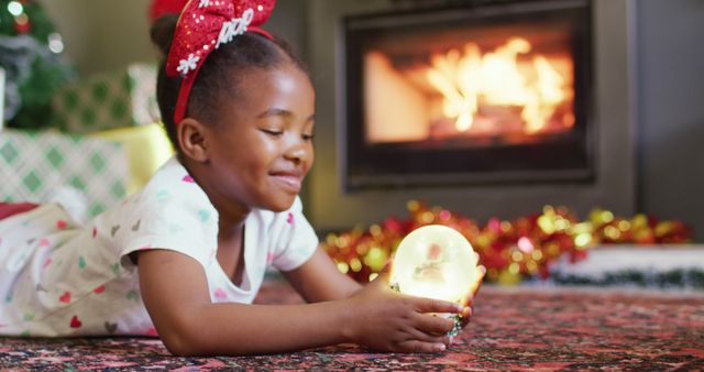 A cheerful child is lying on the floor, holding a snow globe and smiling contentedly near a cozy fireplace. The room is adorned with festive decorations and presents, capturing the warmth and joy of the holiday season. Ideal for use in holiday greeting cards, advertisements, or social media posts conveying warmth, family, and celebration during Christmas.
