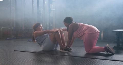 Assisted sit-ups by two women on mat in gym, fitness partners training core together
