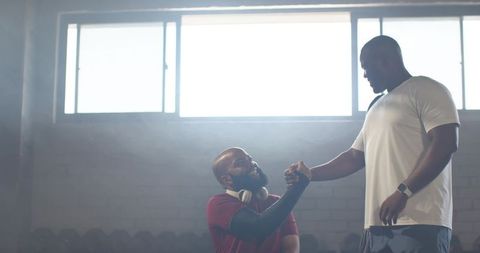African american coach helping athlete up in industrial gym after intense strength training
