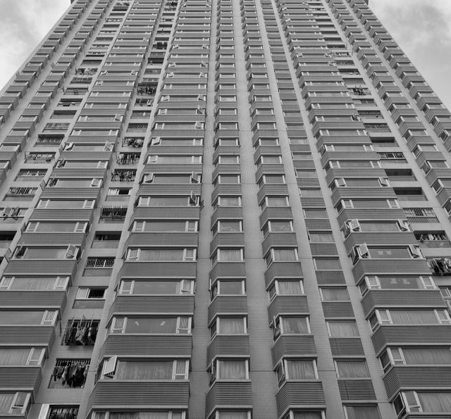 This stock photo depicts the facade of a high-rise apartment building shot from a low angle in black and white. The repetitive pattern of windows gives a sense of the building's scale. This photo can be used for urban development presentations, architectural studies, real estate marketing, or as a representative image of modern city living.
