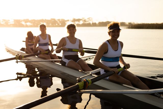 Four Caucasian women rowing team training on a river at sunset, smiling at the camera. Ideal for use in sports and fitness promotions, teamwork and collaboration themes, outdoor activity advertisements, and motivational content.