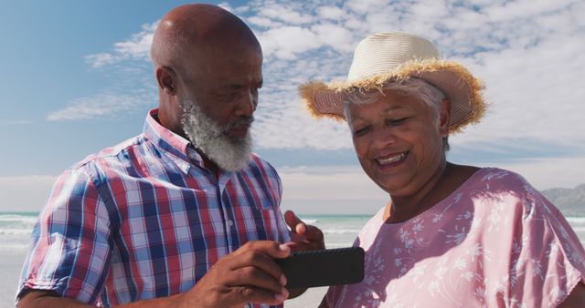 Senior couple enjoying beach while using smartphone, smiling and laughing together. Perfect for vacation advertisements, retired lifestyle promotions, outdoor activities, senior technology use, and travel brochures.
