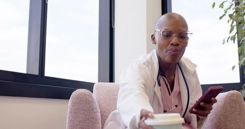 Clinician reaching for coffee in staff lounge holding smartphone and stethoscope in scrubs