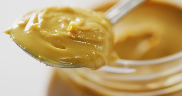 Close-up view of fresh, creamy peanut butter spread on metal spoon with a jar beneath. Smooth texture highlighted in natural lighting. Ideal for illustrating food blogs, nutrition articles, or advertisements promoting high-protein or snack foods.