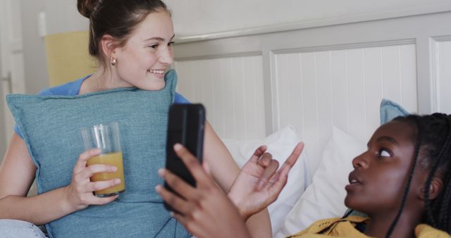 Two teenage friends are relaxing indoors, engaging in a friendly conversation with a smartphone in hand. One holds a glass of orange juice and smiles while the other interacts. This image captures a cozy moment of bonding and can be used in articles or advertisements focused on friendship, youth leisure activities, and at-home relaxation.